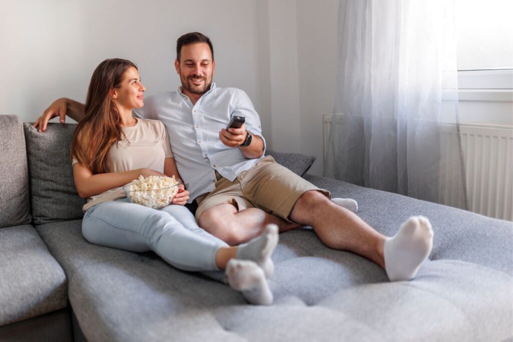 Selecionamos uma imagem simbólica para representar um casal gestante em busca de filmes inspiradores para esta nova fase da vida, para podermos escrever um pouquinho sobre 10 filmes imperdíveis. Na imagem, aparece um casal que recém descobriu a gravidez, a gestante está segurando um pote de pipoca e o pai o controle remoto da TV.